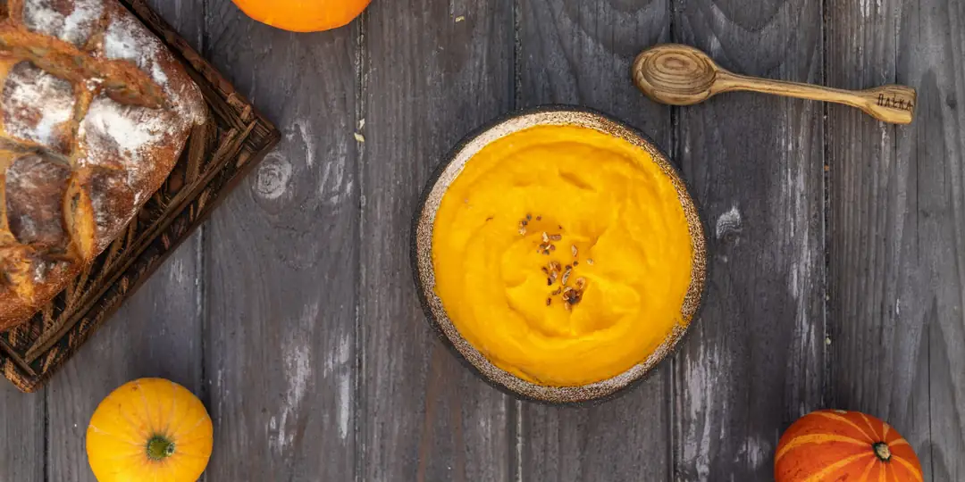 Creme de abóbora amarelo numa tigela sobre a mesa de madeira, com pão e abóboras pequenas