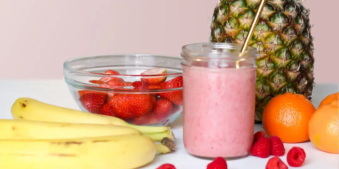 Vitamina rosa de morango e banana num pote de vidro, com bananas, morangos e abacaxi em volta